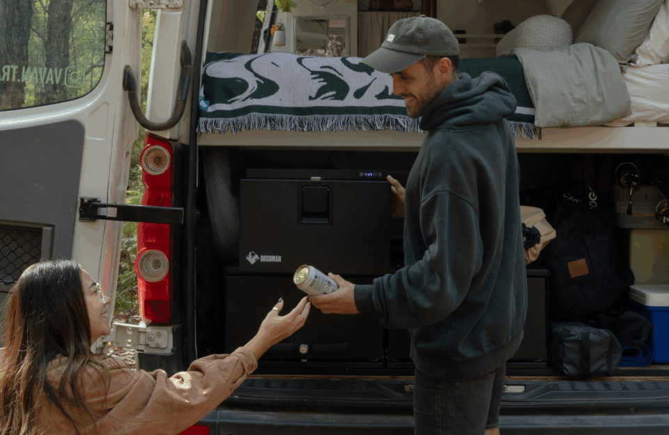 Vee & Andreas at the open rear of the van, handing a cold can between them — the Bushman drawer fridge
