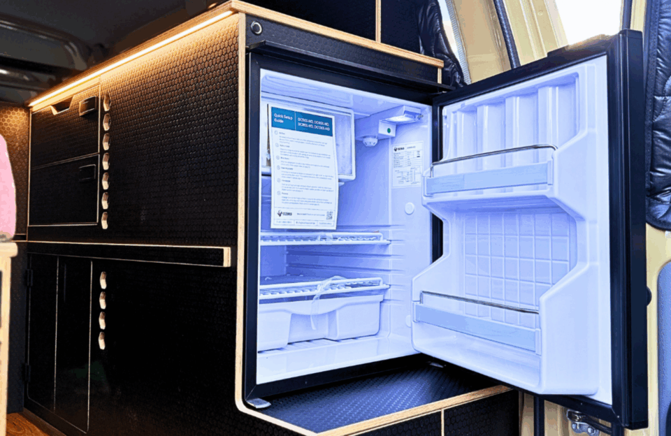 Bushman DC-X Series 12V upright compressor fridge with door open, showing empty interior shelves and quick setup guide, built into black hexagon-panel timber cabinetry inside a campervan fitout.